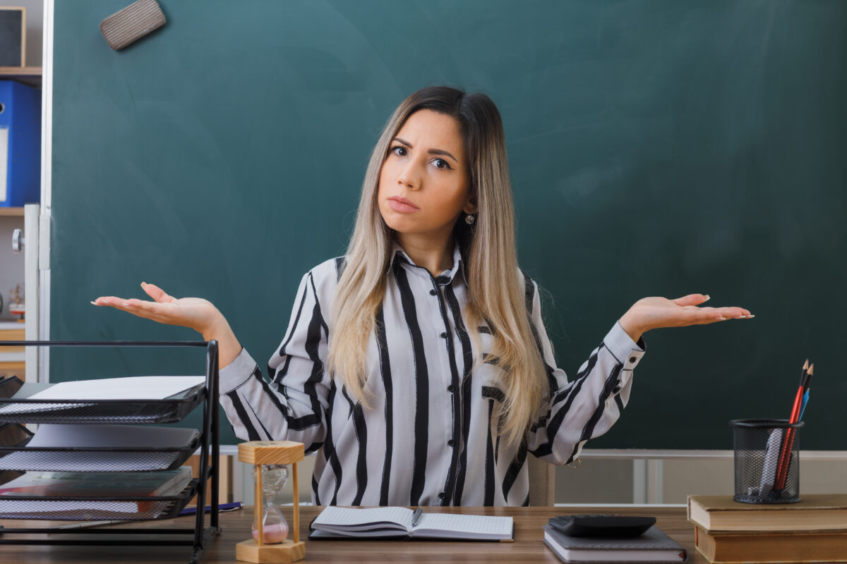 young woman teacher sitting school desk front blackboard classroom checking homework students looking disappointed spreading arms sides